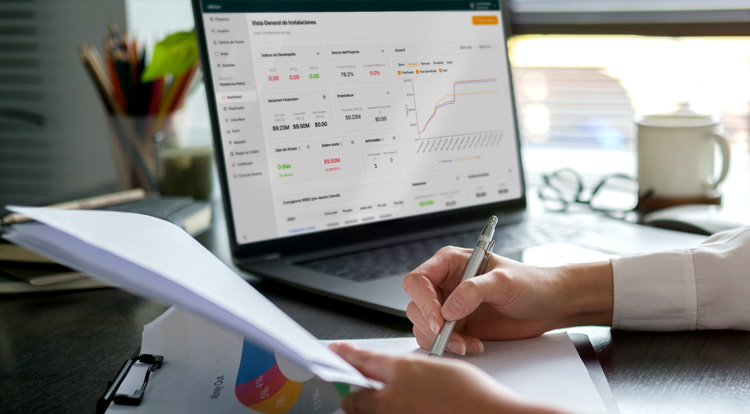 Persona revisando documentos y tomando notas frente a una laptop que muestra un panel de control financiero con gráficos y estadísticas.